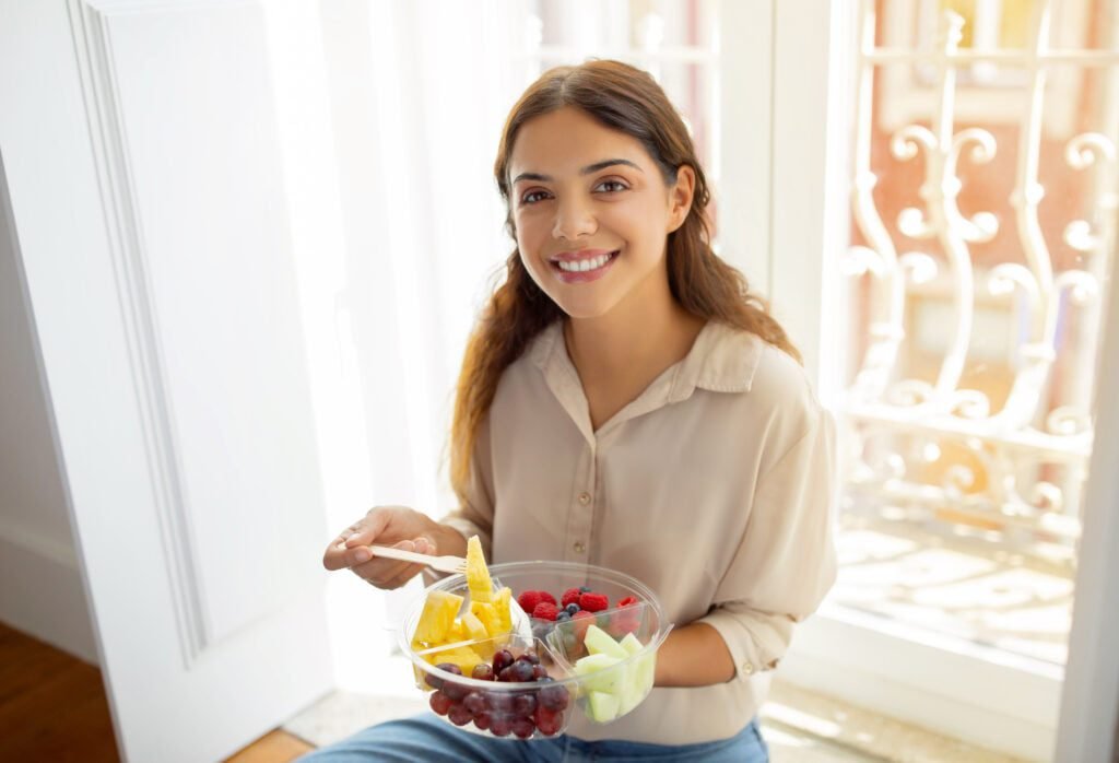 Happy woman holding bowl of fruits enjoying a healthy snack by window